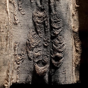 May include: Close-up of distressed fabric with a dark gray hue. The fabric features multiple, large, frayed openings, revealing the underlying texture. The edges of the openings are frayed, creating a textured, worn appearance. The overall effect is a vintage, distressed look.