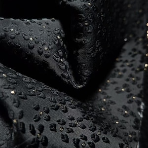 Puede incluir: Una tela negra que presenta un patrón floral en relieve y texturizado en toda la superficie. El material muestra un brillo sutil, con pequeños acentos metálicos de tono dorado incrustados en el centro de cada motivo floral individual.