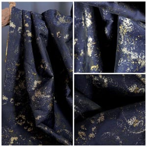 May include: Close-up of a navy blue fabric with gold flecks. The fabric is draped, showing folds and texture. The gold accents create a speckled pattern across the dark blue material, adding a touch of elegance.