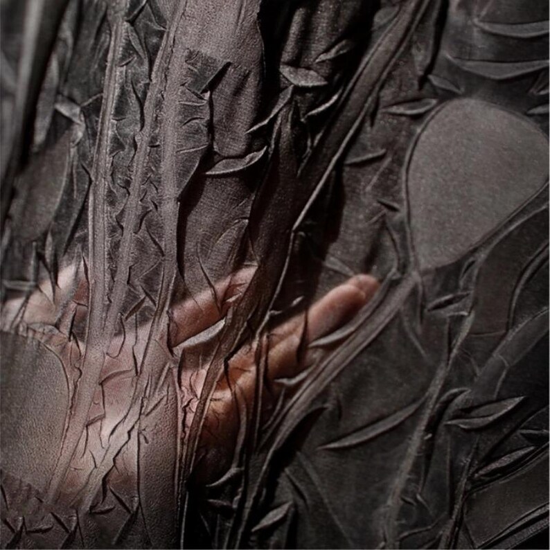 May include: Close-up of black fabric with a textured, crinkled surface. The fabric has a pattern of raised, leaf-like shapes and a sheer quality. The hand is visible through the material, highlighting the fabric's transparency.