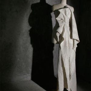 May include: A cream-colored, draped gown on a mannequin. The dress features an asymmetrical design with a one-shoulder detail and a flowing skirt with a high slit. The fabric appears textured, and the dress is displayed against a dark gray background.