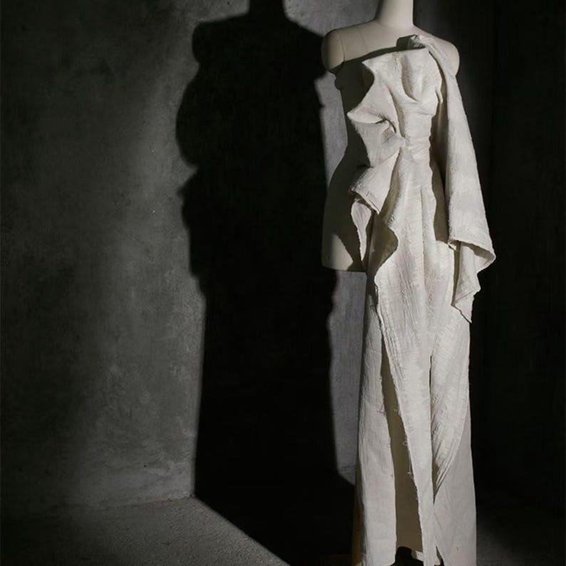 May include: A cream-colored, draped gown on a mannequin. The dress features an asymmetrical design with a one-shoulder detail and a flowing skirt with a high slit. The fabric appears textured, and the dress is displayed against a dark gray background.