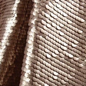 May include: A fabric textile covered in small, round, metallic champagne-colored sequins arranged in a dense, overlapping pattern. The surface reflects light, creating highlights and shadows across the curved folds of the material.