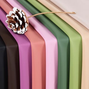 May include: A close-up of a variety of colourful, folded sheets of paper. The colours include black, brown, purple, coral, pink, green, and beige. A pine cone with white accents rests on top of the sheets, connected by a thin, brown string.