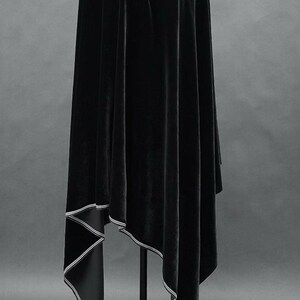 May include: A draped black velvet garment with a contrasting white trim. The fabric hangs in soft folds, creating a flowing silhouette. The hemline features a geometric design with a white border. The garment is displayed on a black stand against a gray backdrop.