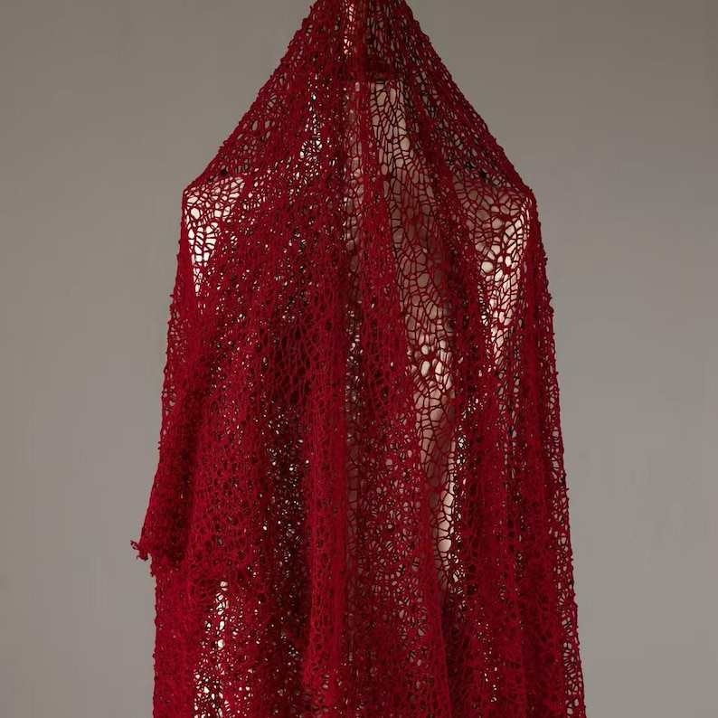 May include: A draped, open-weave textile in a deep red hue. The fabric is suspended, creating a flowing, textured effect. The intricate pattern of the weave is visible, adding depth and visual interest to the piece. The textile appears to be lightweight.