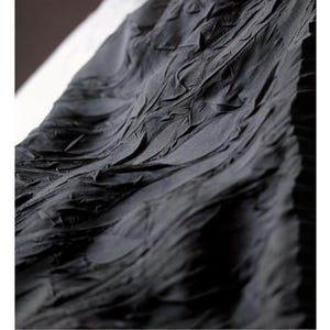 May include: Close-up of a dark gray fabric with a textured, rippled surface. The fabric appears to be draped, with folds and ridges creating a dynamic pattern. The material has a matte finish, and the lighting highlights the texture.