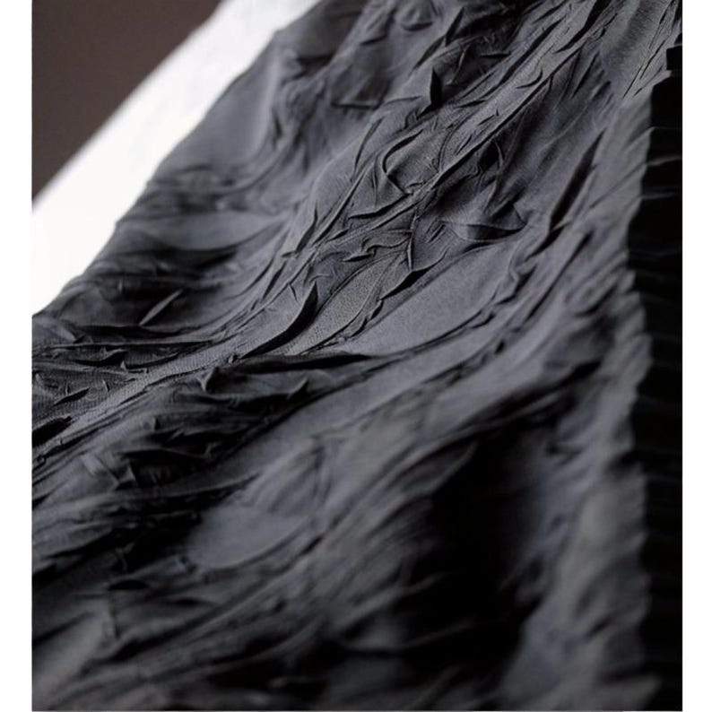May include: Close-up of a dark gray fabric with a textured, rippled surface. The fabric appears to be draped, with folds and ridges creating a dynamic pattern. The material has a matte finish, and the lighting highlights the texture.