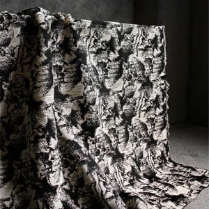 以下が含まれることがあります： 黒と白の模様の生地が表面にかけられています。生地は、繰り返しのデザインでテクスチャ加工された外観をしています。パターンは抽象的な形と線の混合であり、視覚的に面白いテキスタイルを作成しています。