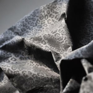 Puede incluir: Primer plano de una tela con estampado de leopardo. La tela es de color gris oscuro con manchas grises más claras. La tela está doblada y drapeada, creando sombras y textura.