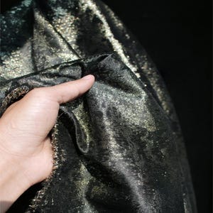 Puede incluir: Primer plano de una tela negra con detalles de purpurina dorada. La tela se sostiene, mostrando su textura y brillo. El material parece suave y fluido, adecuado para prendas de vestir o proyectos decorativos.