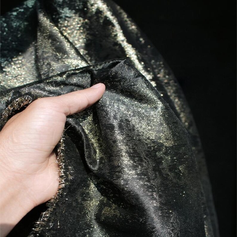Puede incluir: Primer plano de una tela negra con detalles de purpurina dorada. La tela se sostiene, mostrando su textura y brillo. El material parece suave y fluido, adecuado para prendas de vestir o proyectos decorativos.