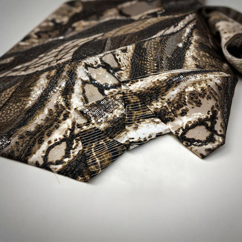 May include: A patterned necktie in shades of brown, beige, and black. The tie features a repeating design with a mix of textures, including sequins. The tie is folded over, showing the intricate pattern.