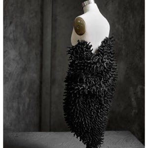 May include: A black, textured garment displayed on a white mannequin. The garment has a dense, spiky appearance, resembling a mass of dark, irregular protrusions. The mannequin is set against a gray, textured background, highlighting the garment's unique design.