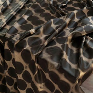 May include: Close-up of a fabric with a repeating pattern of dark brown, oval shapes against a tan background. The fabric is draped, showing folds and shadows that add depth and texture. The overall effect is a blend of organic and geometric design.