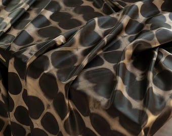 Puede incluir: Primer plano de una tela con un patrón repetitivo de formas ovaladas de color marrón oscuro sobre un fondo beige. La tela está drapeada, mostrando pliegues y sombras que añaden profundidad y textura. El diseño combina elementos orgánicos y geométricos.