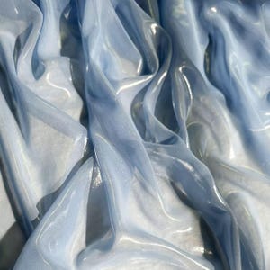 Könnte beinhalten: Nahaufnahme von fließendem, durchscheinendem Stoff in Hellblau- und Weißtönen. Der Stoff hat eine schimmernde, irisierende Qualität mit weichen Falten und Glanzlichtern. Das Material wirkt zart und luftig.