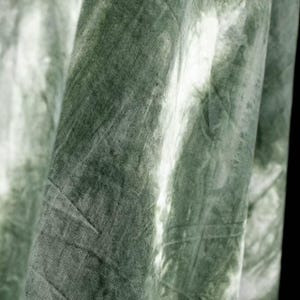 Puede incluir: Primer plano de una tela con efecto tie-dye. La tela es de color verde apagado con zonas m&aacute;s claras, casi blancas, creando una apariencia marmolada. El material parece suave y tiene pliegues y texturas visibles.