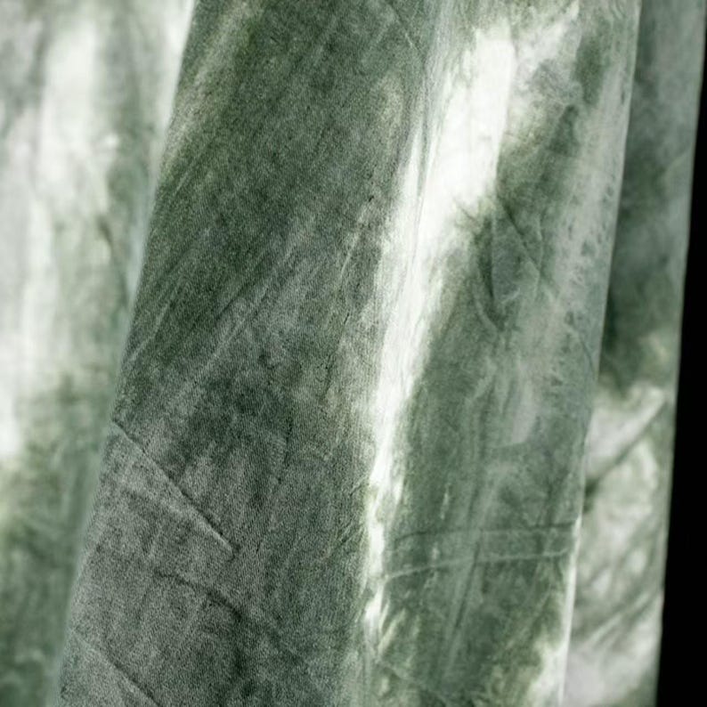 Puede incluir: Primer plano de una tela con efecto tie-dye. La tela es de color verde apagado con zonas m&aacute;s claras, casi blancas, creando una apariencia marmolada. El material parece suave y tiene pliegues y texturas visibles.