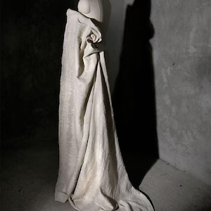 May include: A cream-colored fabric draped over a mannequin. The fabric has a subtle, textured pattern and hangs in soft folds. The lighting creates shadows that emphasize the fabric's texture and shape. The background is a dark, textured wall.