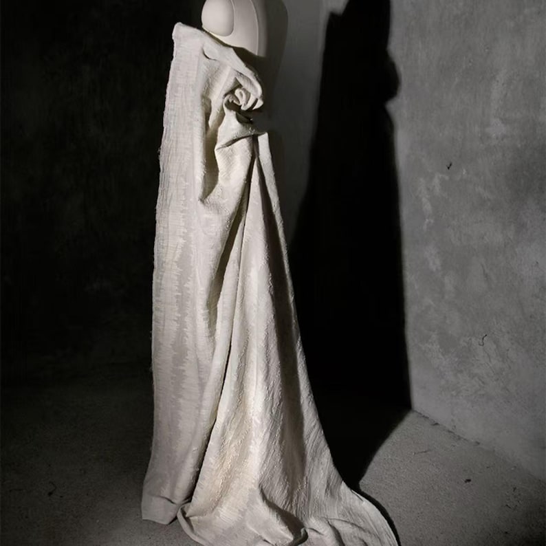 May include: A cream-colored fabric draped over a mannequin. The fabric has a subtle, textured pattern and hangs in soft folds. The lighting creates shadows that emphasize the fabric's texture and shape. The background is a dark, textured wall.