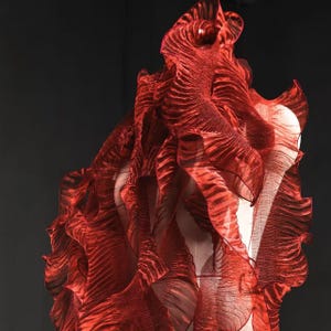 以下が含まれることがあります： 鮮やかな赤色のフリル付き衣服で、流れるようなテクスチャードデザイン。生地は透けており、波状のパターンがあり、ダイナミックでレイヤー状の効果を生み出しています。マネキンに展示されており、ファッションやオートクチュール作品を示唆しています。