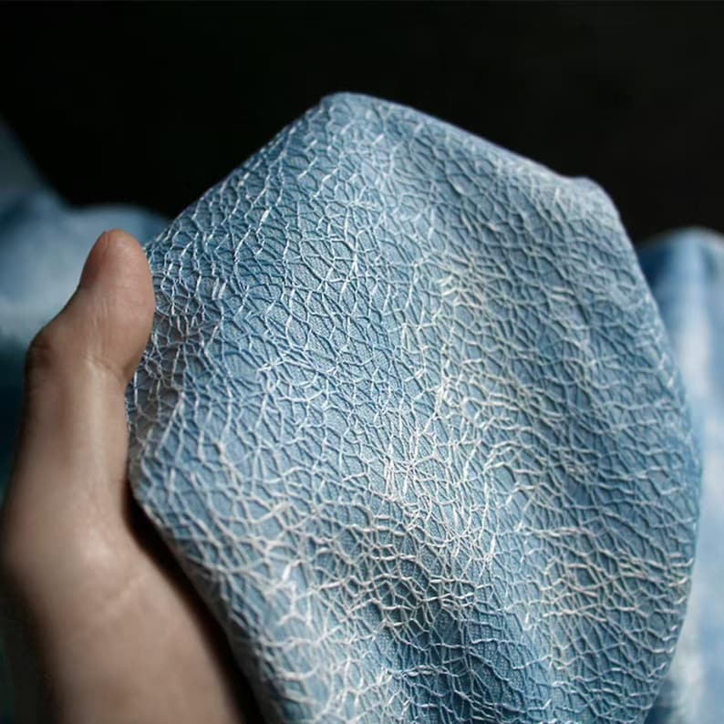 Op de afbeelding: Close-up van blauwe stof met een textuurpatroon in de vorm van een wit web. De stof wordt in een hand gehouden, wat het gedetailleerde ontwerp benadrukt. De afbeelding richt zich op de textuur en kleur van het materiaal.