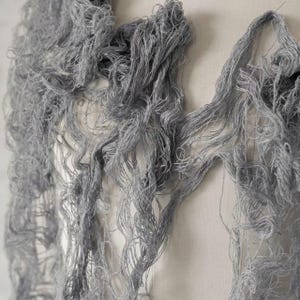 May include: A light gray textile piece featuring a loose, open-weave construction with long, dangling, frayed threads. The material has a distressed, fibrous appearance with a tangled, organic texture that creates a layered, semi-sheer effect.
