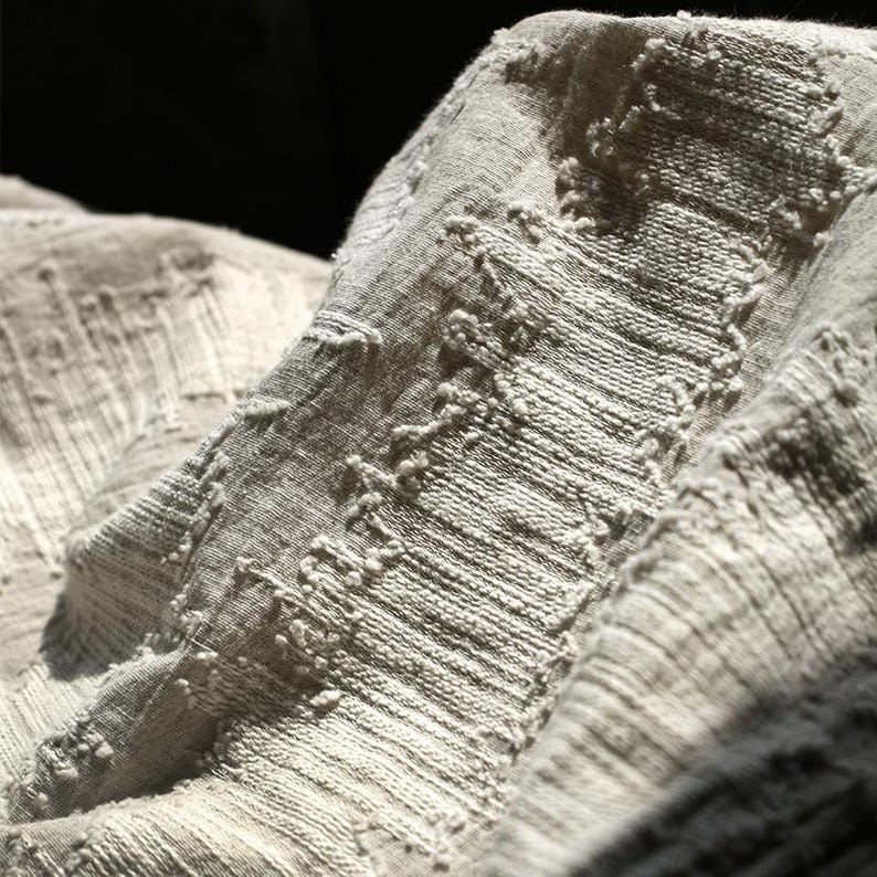May include: Close-up of a textured, light gray fabric with a woven pattern. The fabric has a slightly wrinkled appearance, with raised, irregular stitching creating a tactile surface. The lighting highlights the texture and depth of the material.