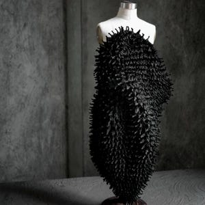 May include: A black, textured garment on a white dress form. The garment has a dense, spiky texture, creating a unique visual effect. The dress form is set against a gray background, highlighting the garment's form and texture. The overall aesthetic is modern and artistic.