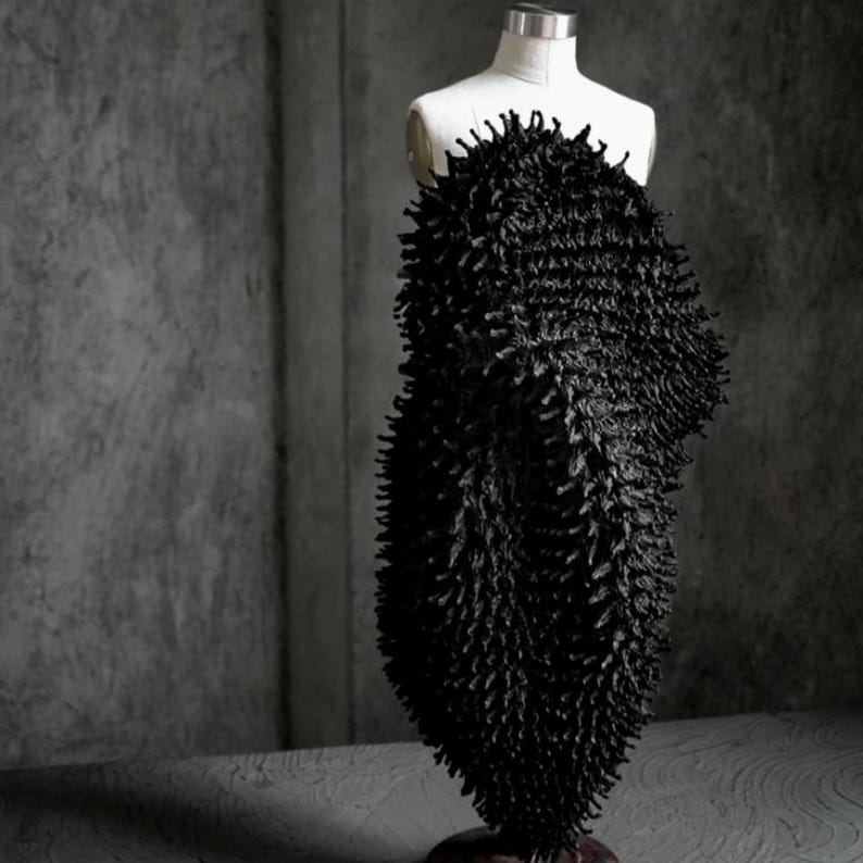 May include: A black, textured garment on a white dress form. The garment has a dense, spiky texture, creating a unique visual effect. The dress form is set against a gray background, highlighting the garment's form and texture. The overall aesthetic is modern and artistic.
