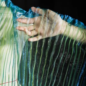 May include: A sheer, pleated fabric in shades of blue and green. The fabric is draped, with vertical lines creating a textured effect. The fabric is held by a hand, with the fingers visible through the translucent material.
