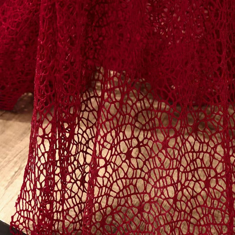 May include: Close-up of a deep red, open-weave fabric. The material has a delicate, net-like pattern with a slightly textured appearance. The fabric is draped, creating shadows and highlighting the intricate design. The overall effect is elegant and airy.