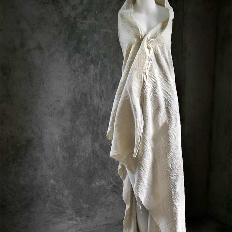 May include: A cream-colored, draped garment on a mannequin. The fabric has a textured appearance and is wrapped around the form, creating soft folds and a hooded neckline. The background is a mottled gray.
