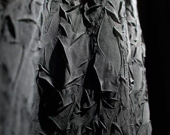 May include: Close-up of a dark gray fabric with a textured, layered design. The fabric appears to have a three-dimensional quality, with irregular shapes and folds creating shadows and highlights. The overall effect is dramatic and artistic.