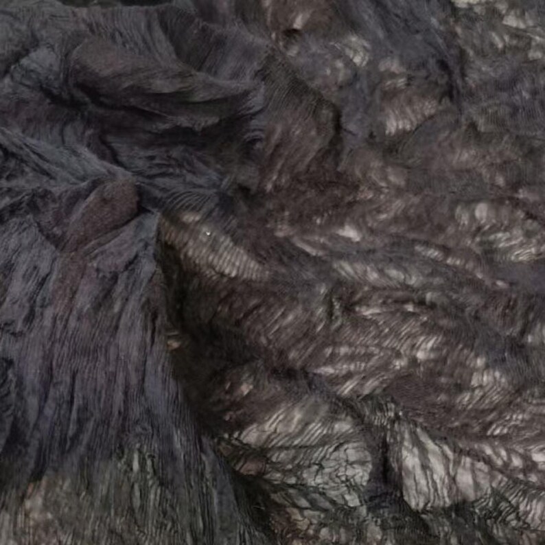 May include: A close-up view of a dark, textured fabric with a layered appearance. The fabric has a mix of dark gray and black tones, creating a shadowed effect. The material appears soft and flowing, with visible folds and ridges.