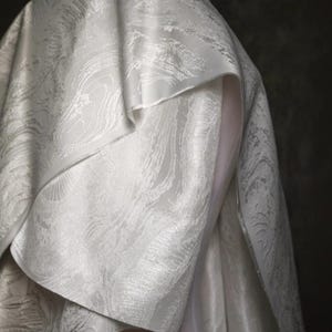K&ouml;nnte beinhalten: Nahaufnahme eines wei&szlig;en Stoffes mit einem strukturierten, marmorierten Muster. Der Stoff ist drapiert, wobei Falten und Schichten sichtbar sind. Das Material wirkt dick und luxuri&ouml;s, mit einem dezenten Glanz. Der Hintergrund ist dunkel und neutral.