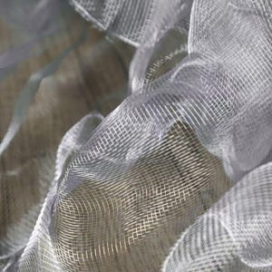 May include: Close-up of a sheer, light gray fabric with a grid-like pattern. The fabric is draped and folded, creating shadows and highlights. The material appears delicate and possibly used for decorative purposes.