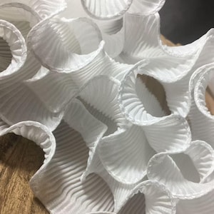 May include: Close-up of white, ruffled fabric with a pleated texture. The fabric is arranged in overlapping, swirling patterns, creating a layered effect. The edges of the fabric are softly curved, and the overall appearance is delicate and airy.