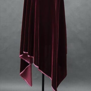 May include: A draped, burgundy velvet fabric with a contrasting pink trim. The fabric hangs in soft folds, creating a sense of movement. The material appears to be thick and luxurious, with a subtle sheen.
