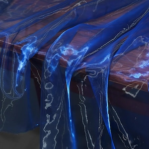 Pode incluir: Close-up de tecido azul transparente e fluido com um efeito brilhante subtil. O tecido drapeia sobre uma superfície escura, criando um jogo de luz e sombra. O material tem uma qualidade delicada, quase etérea, com finos padrões brancos.