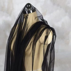 Puede incluir: Tela de tul negra drapeada sobre un maniquí. El material transparente está en capas, creando un efecto fluido. La tela es opaca en algunas áreas y translúcida en otras, revelando el maniquí debajo. El fondo es de un color claro y neutro.