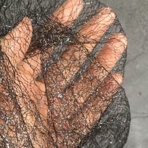 Könnte beinhalten: Nahaufnahme von schwarzem Netz mit silbernen Metallicfäden. Das Netz wird hochgehalten, wodurch ein strukturiertes, netzartiges Aussehen entsteht. Das Material wird wahrscheinlich für dekorative Zwecke oder zum Basteln verwendet.