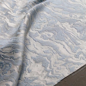 Puede incluir: Primer plano de una tela doblada con un diseño veteado. La tela presenta una mezcla de tonos azul claro y blanco, creando una apariencia fluida y texturizada. El material parece ser un textil lujoso, posiblemente para tapicería o cortinas.