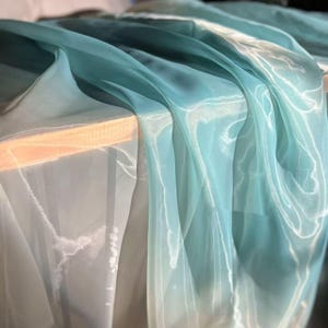 Peut inclure: Gros plan sur un tissu fluide et transparent dans des tons de bleu sarcelle et de blanc. Le tissu est drapé sur une surface en bois, créant des plis et des reflets doux. Le matériau semble léger et translucide, avec un éclat subtil.
