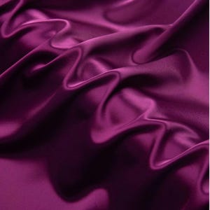 May include: A deep magenta fabric with a smooth, lustrous finish. The material is draped in soft, flowing folds that create a series of highlights and shadows across the surface, emphasising the reflective quality of the textile's texture.