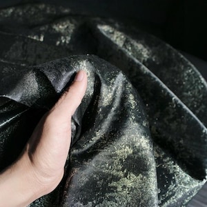 Puede incluir: Tela negra con un patr&oacute;n dorado met&aacute;lico. El material est&aacute; drapeado, creando pliegues y sombras. La tela tiene un brillo sutil y los acentos dorados a&ntilde;aden un toque de lujo. La textura general parece suave y fluida.