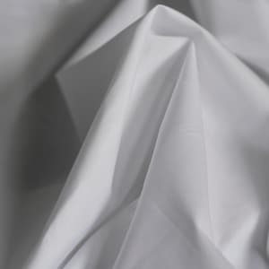 Pode incluir: Close-up de tecido branco com dobras suaves e sombras. O tecido parece leve e fluido, com um brilho sutil. A imagem destaca a textura e a queda do material.