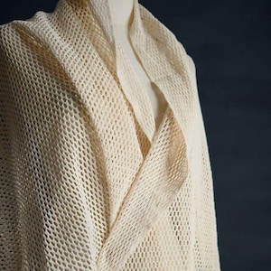 May include: Cream-colored, open-weave shawl draped over a mannequin. The shawl has a loose, net-like texture with a draped collar. The fabric appears soft and lightweight, suitable for layering. The background is a dark, solid color.