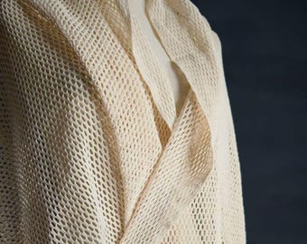 May include: Cream-colored, open-weave shawl draped over a mannequin. The shawl has a loose, net-like texture with a draped collar. The fabric appears soft and lightweight, suitable for layering. The background is a dark, solid color.